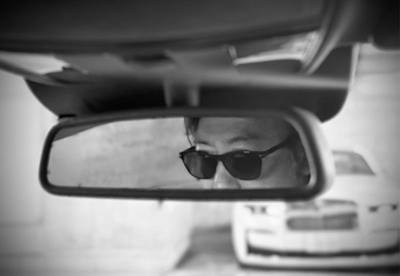 車のルームミラーに映るサングラス姿の西田理一郎（モノクロ）